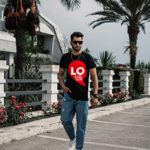 Puede incluir: Una camiseta negra de manga corta con un gr&aacute;fico en forma de coraz&oacute;n rojo en el pecho. El gr&aacute;fico incluye el texto blanco LO y el texto m&aacute;s peque&ntilde;o R U MY OTHER HALF? La camiseta se combina con pantalones vaqueros azules y zapatillas blancas.