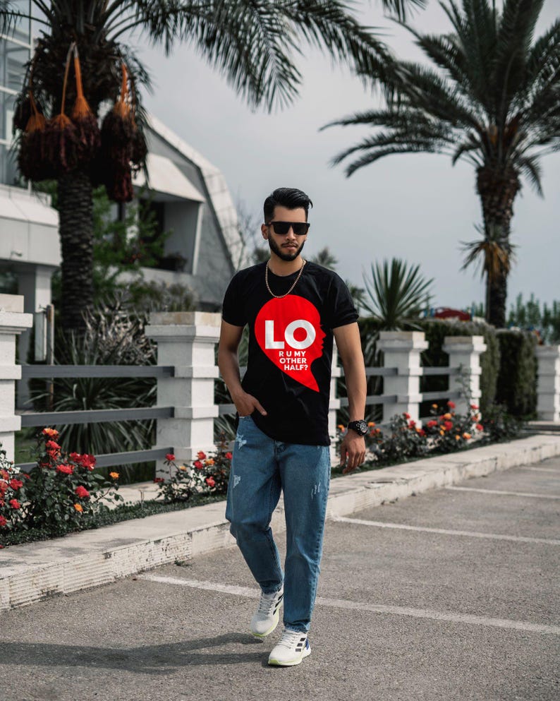 Puede incluir: Una camiseta negra de manga corta con un gr&aacute;fico en forma de coraz&oacute;n rojo en el pecho. El gr&aacute;fico incluye el texto blanco LO y el texto m&aacute;s peque&ntilde;o R U MY OTHER HALF? La camiseta se combina con pantalones vaqueros azules y zapatillas blancas.