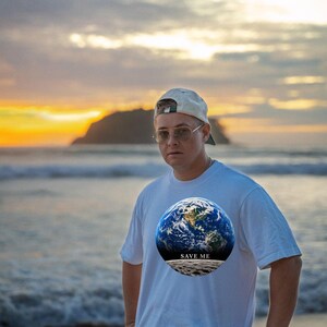 May include: A white short-sleeved t-shirt featuring a circular graphic print of the Earth above a lunar surface with the text SAVE ME printed in black. The shirt is worn with a light-colored baseball cap and metal-framed glasses.