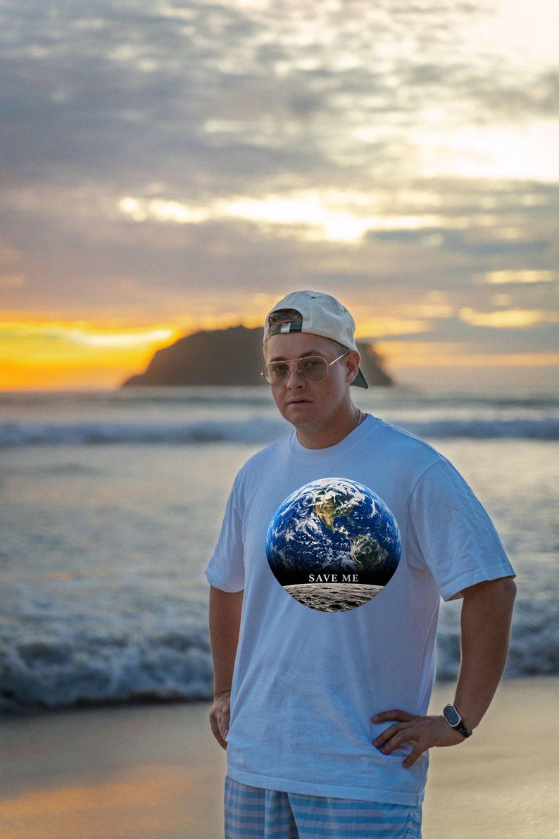 May include: A white short-sleeved t-shirt featuring a circular graphic print of the Earth above a lunar surface with the text SAVE ME printed in black. The shirt is worn with a light-colored baseball cap and metal-framed glasses.