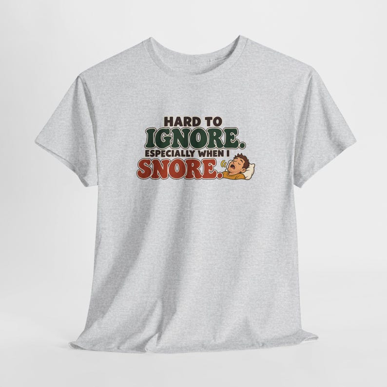 May include: A light gray short-sleeve t-shirt featuring the text "HARD TO IGNORE. ESPECIALLY WHEN I SNORE." in dark green and red lettering, accompanied by a cartoon illustration of a person sleeping and snoring on a white pillow.