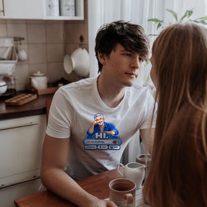 May include: A white short-sleeved t-shirt featuring a graphic print of a person resting their chin on their hand above the text HI. and Ask me anything... with icons for chat, voice, and image, set against a blue background.