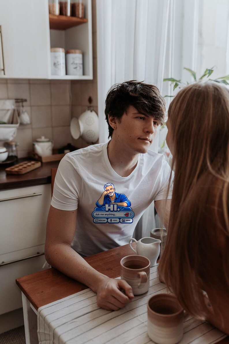 May include: A white short-sleeved t-shirt featuring a graphic print of a person resting their chin on their hand above the text HI. and Ask me anything... with icons for chat, voice, and image, set against a blue background.