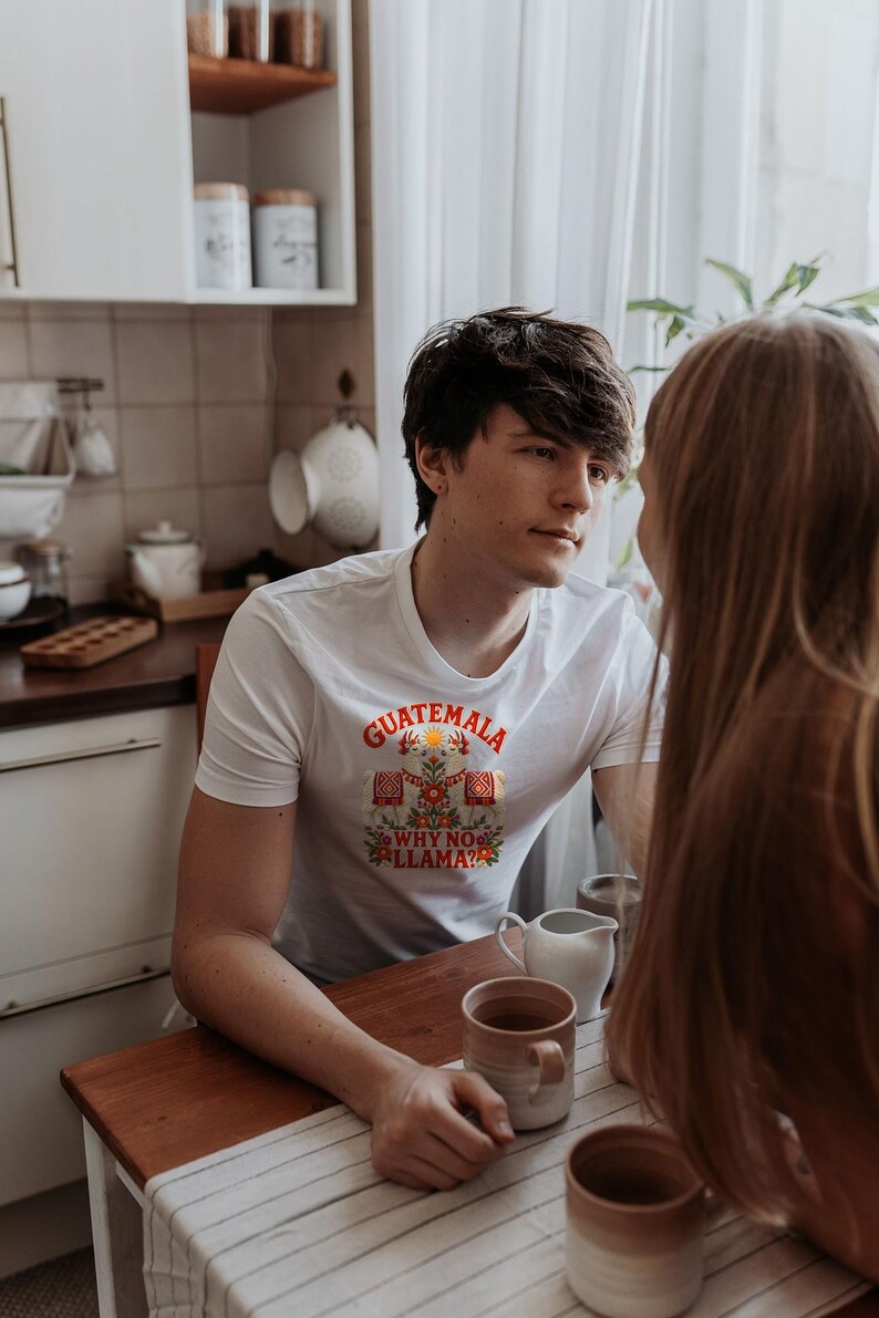 Puede incluir: Una camiseta blanca de manga corta con un estampado gr&aacute;fico colorido de dos llamas, motivos florales y el texto "GUATEMALA" arriba y "WHY NO LLAMA?" debajo de la ilustraci&oacute;n, centrado en el pecho de la prenda.
