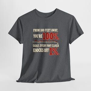 May include: A charcoal gray short-sleeve t-shirt featuring the text: FROM 100 FEET AWAY, YOU'RE 100%. SADLY, EVERY FOOT CLOSER KNOCKS OFF 1%. The design includes a horizontal line graph with small red marks and the numbers 100% and 1% in red.