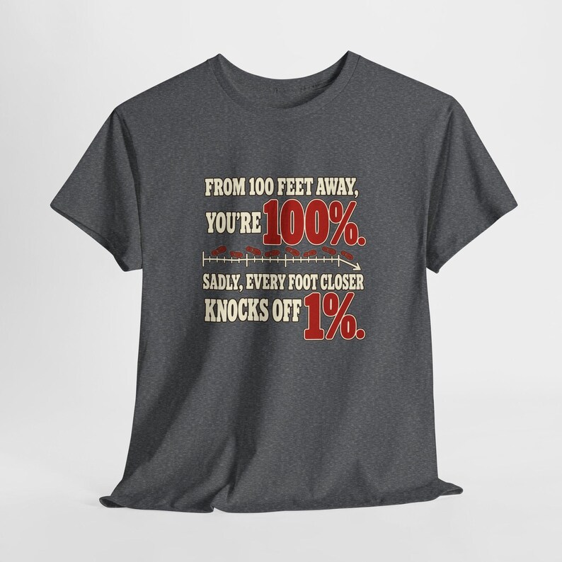 May include: A charcoal gray short-sleeve t-shirt featuring the text: FROM 100 FEET AWAY, YOU'RE 100%. SADLY, EVERY FOOT CLOSER KNOCKS OFF 1%. The design includes a horizontal line graph with small red marks and the numbers 100% and 1% in red.