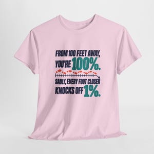 May include: A light pink short-sleeve t-shirt featuring dark blue and teal text that reads: FROM 100 FEET AWAY, YOU'RE 100%. SADLY, EVERY FOOT CLOSER KNOCKS OFF 1%. The design includes a horizontal measurement line with small red footprints above it.