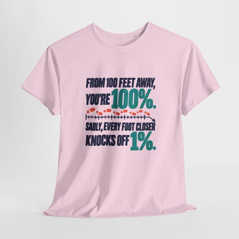 May include: A light pink short-sleeve t-shirt featuring dark blue and teal text that reads: FROM 100 FEET AWAY, YOU'RE 100%. SADLY, EVERY FOOT CLOSER KNOCKS OFF 1%. The design includes a horizontal measurement line with small red footprints above it.