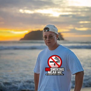 May include: A white short-sleeved t-shirt featuring a red circular no-smoking symbol with a cigarette and a small figure. Below the graphic, the text reads NO SMOKING NEAR ME and MY PERSONAL SPACE IS SMOKE-FREE in bold black and red lettering.