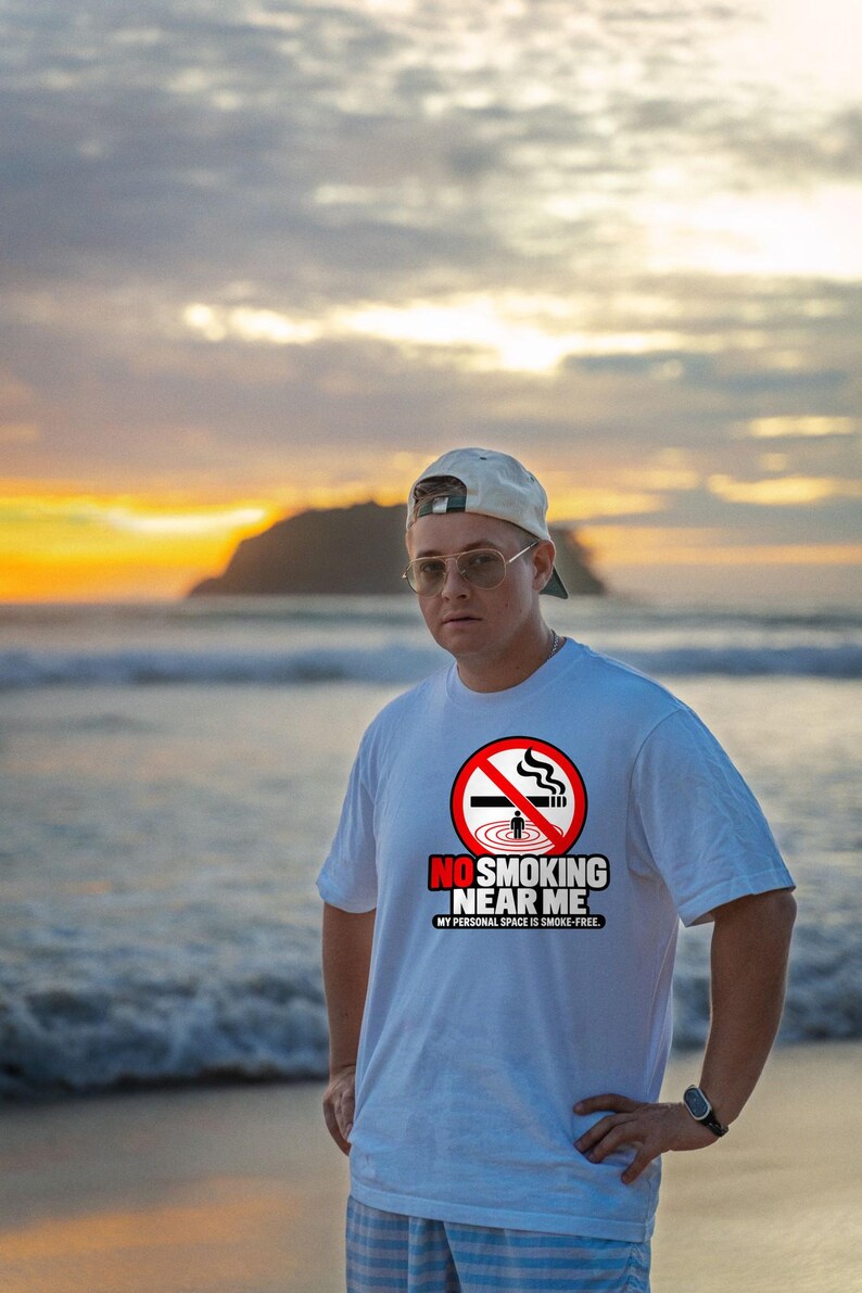 May include: A white short-sleeved t-shirt featuring a red circular no-smoking symbol with a cigarette and a small figure. Below the graphic, the text reads NO SMOKING NEAR ME and MY PERSONAL SPACE IS SMOKE-FREE in bold black and red lettering.