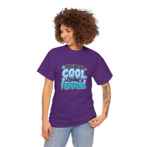 May include: A purple short-sleeve t-shirt featuring a graphic print with the text "YOU'RE SO COOL, I'M FREEZING" in blue, icy, and white lettering, surrounded by snowflake and ice cube illustrations on the front of the garment.