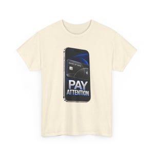 May include: A cream-colored t-shirt featuring a graphic of a smartphone displaying a black credit card labeled BANK OF MINE. Below the card, the text PAY ATTENTION is printed in bold, metallic-toned, capitalized letters.