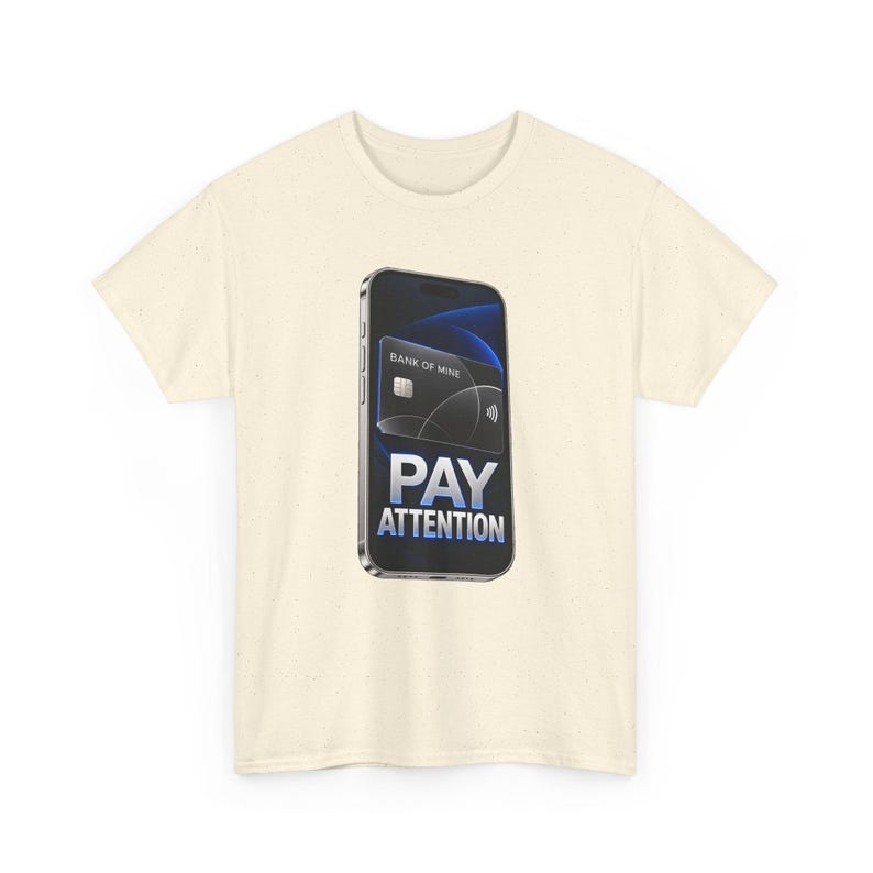 May include: A cream-colored t-shirt featuring a graphic of a smartphone displaying a black credit card labeled BANK OF MINE. Below the card, the text PAY ATTENTION is printed in bold, metallic-toned, capitalized letters.