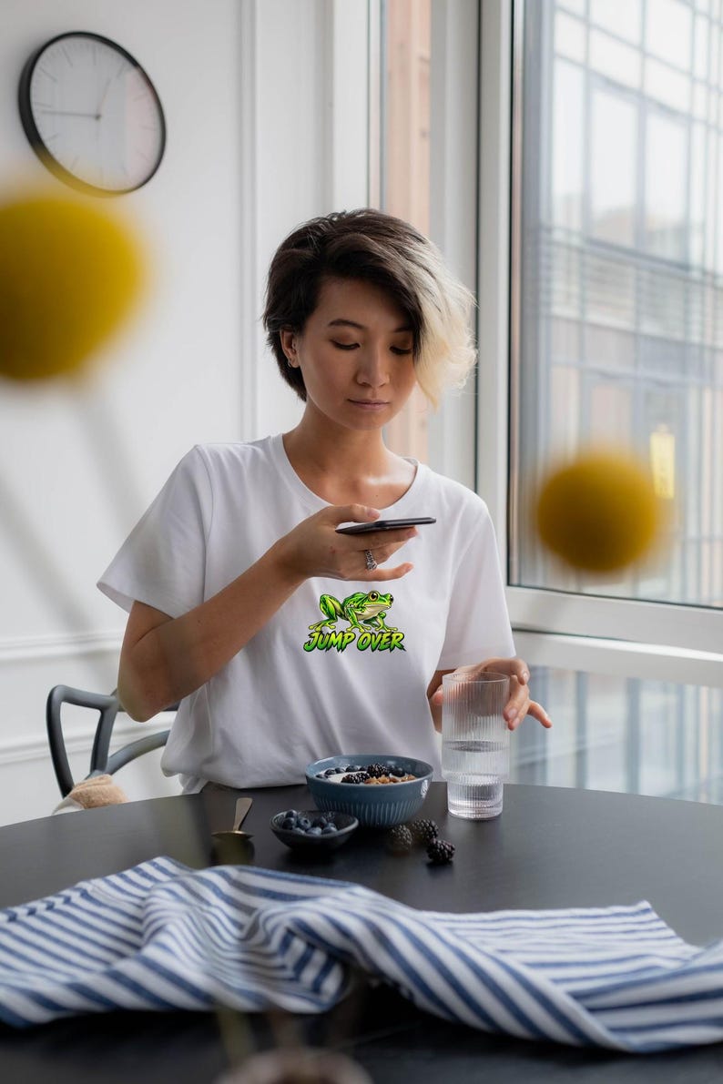 May include: A white short-sleeved t-shirt featuring a graphic of a green frog and the text "JUMP OVER" in a stylized yellow and green font. The shirt has a crew neckline and a relaxed fit, showcasing a clean and simple design.