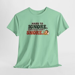 May include: A light green short-sleeve t-shirt featuring the text HARD TO IGNORE. ESPECIALLY WHEN I SNORE. in dark green and red lettering, accompanied by a graphic of a person sleeping on a white pillow with a small sound icon.