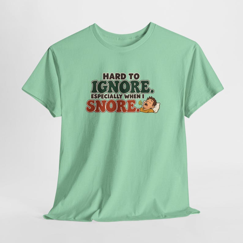 May include: A light green short-sleeve t-shirt featuring the text HARD TO IGNORE. ESPECIALLY WHEN I SNORE. in dark green and red lettering, accompanied by a graphic of a person sleeping on a white pillow with a small sound icon.