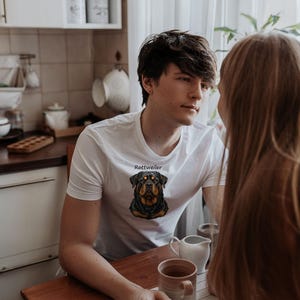 May include: A white short-sleeved t-shirt featuring a graphic illustration of a Rottweiler dog head centered on the chest, with the word Rottweiler printed in black text above the dog's head.