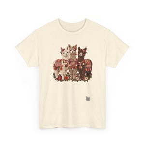 May include: A cream-colored short-sleeve t-shirt featuring a graphic of three llamas in white, tan, and dark brown. Each llama wears a colorful patterned blanket, tassels, and floral headpieces. A small QR code is printed near the bottom right.