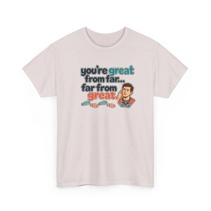 May include: A light pink short-sleeve t-shirt featuring a graphic of a person looking through binoculars with the text "you're great from far... far from great." and small colorful footprints printed on the front center.