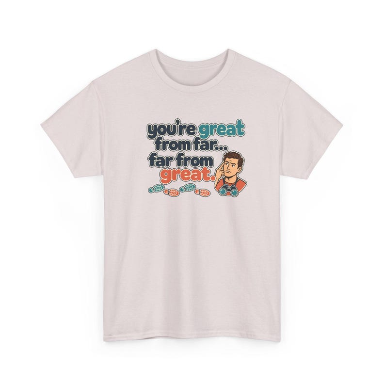 May include: A light pink short-sleeve t-shirt featuring a graphic of a person looking through binoculars with the text "you're great from far... far from great." and small colorful footprints printed on the front center.