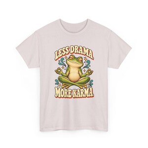 May include: A light pink t-shirt featuring a graphic of a green frog sitting in a lotus position with eyes closed. The text above the frog reads "LESS DRAMA." and the text below reads "MORE KARMA." with small blue water-like accents and stars.