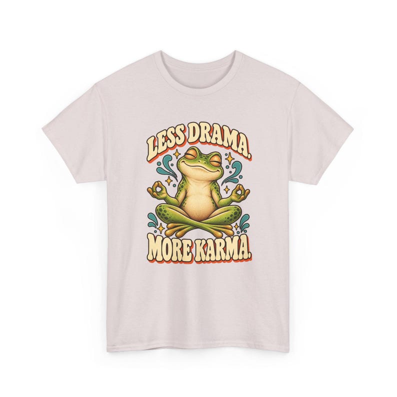 May include: A light pink t-shirt featuring a graphic of a green frog sitting in a lotus position with eyes closed. The text above the frog reads "LESS DRAMA." and the text below reads "MORE KARMA." with small blue water-like accents and stars.