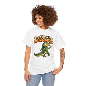 May include: A white short-sleeve t-shirt featuring a graphic of a green alligator waving its hand. Above the alligator, the text reads "SEE YOU LATER ALLIGATOR" in bold, orange, and yellow gradient lettering with a dark outline.