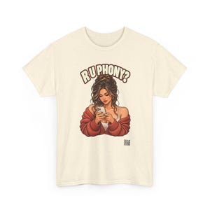 May include: A cream-colored short-sleeve t-shirt featuring a graphic illustration of a person with long brown hair wearing a red cardigan and looking at a smartphone. The text "RU PHONY?" is printed in brown above the illustration.