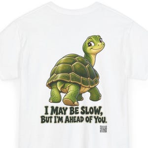 May include: A white t-shirt featuring a cartoon green turtle looking back over its shoulder. Below the turtle, the text reads, "I MAY BE SLOW, BUT I'M AHEAD OF YOU." A small QR code is printed in the bottom right corner of the graphic design.