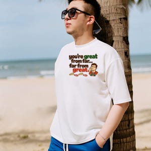May include: A white short-sleeved t-shirt featuring the text "you're great from far... far from great." in brown and green lettering, accompanied by an illustration of a person looking through binoculars and several small footprint icons.