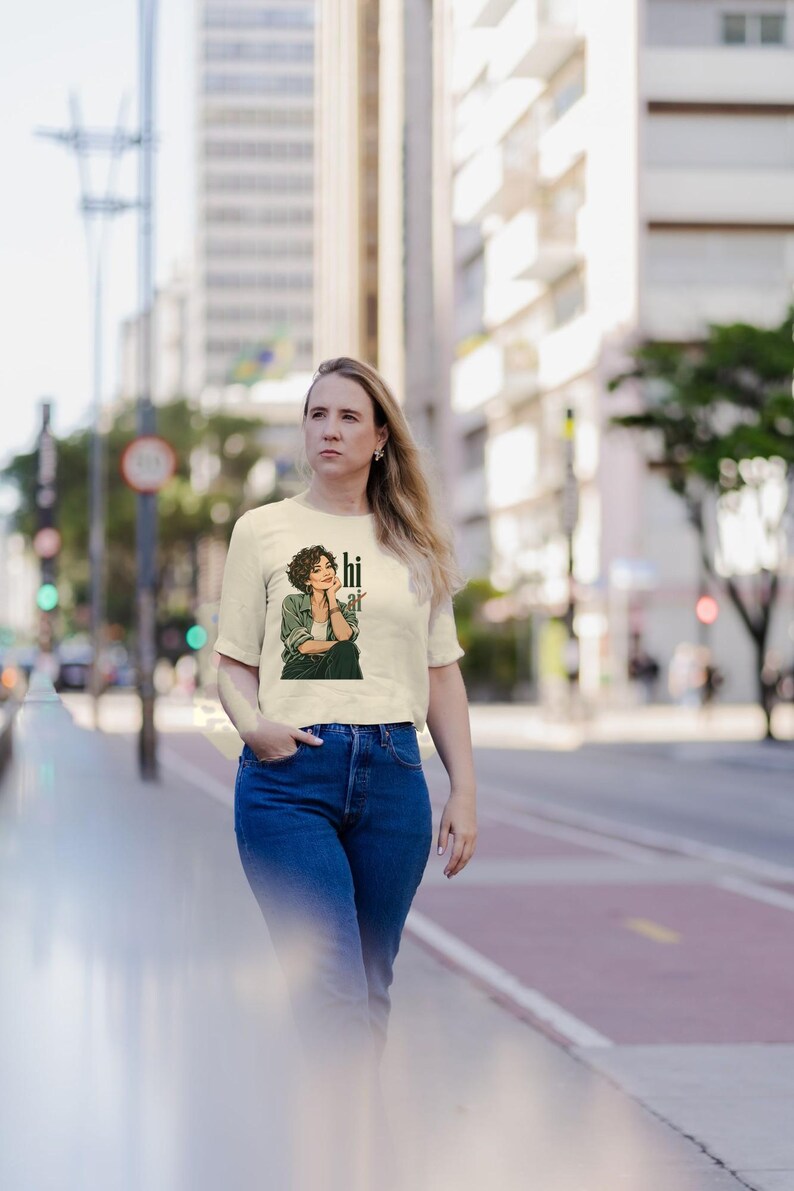 Op de afbeelding: Een cr&egrave;mekleurig T-shirt met korte mouwen en een grafische illustratie van een persoon met krullend haar in een groen overhemd en een donkere broek, met de tekst "hi" verticaal naast de figuur gedrukt in een vet, zwart lettertype.