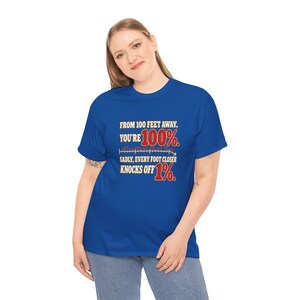 May include: A royal blue short-sleeve t-shirt featuring the text: FROM 100 FEET AWAY, YOU'RE 100%. SADLY, EVERY FOOT CLOSER KNOCKS OFF 1%. The text includes a graphic of a ruler with red numbers and a downward arrow.