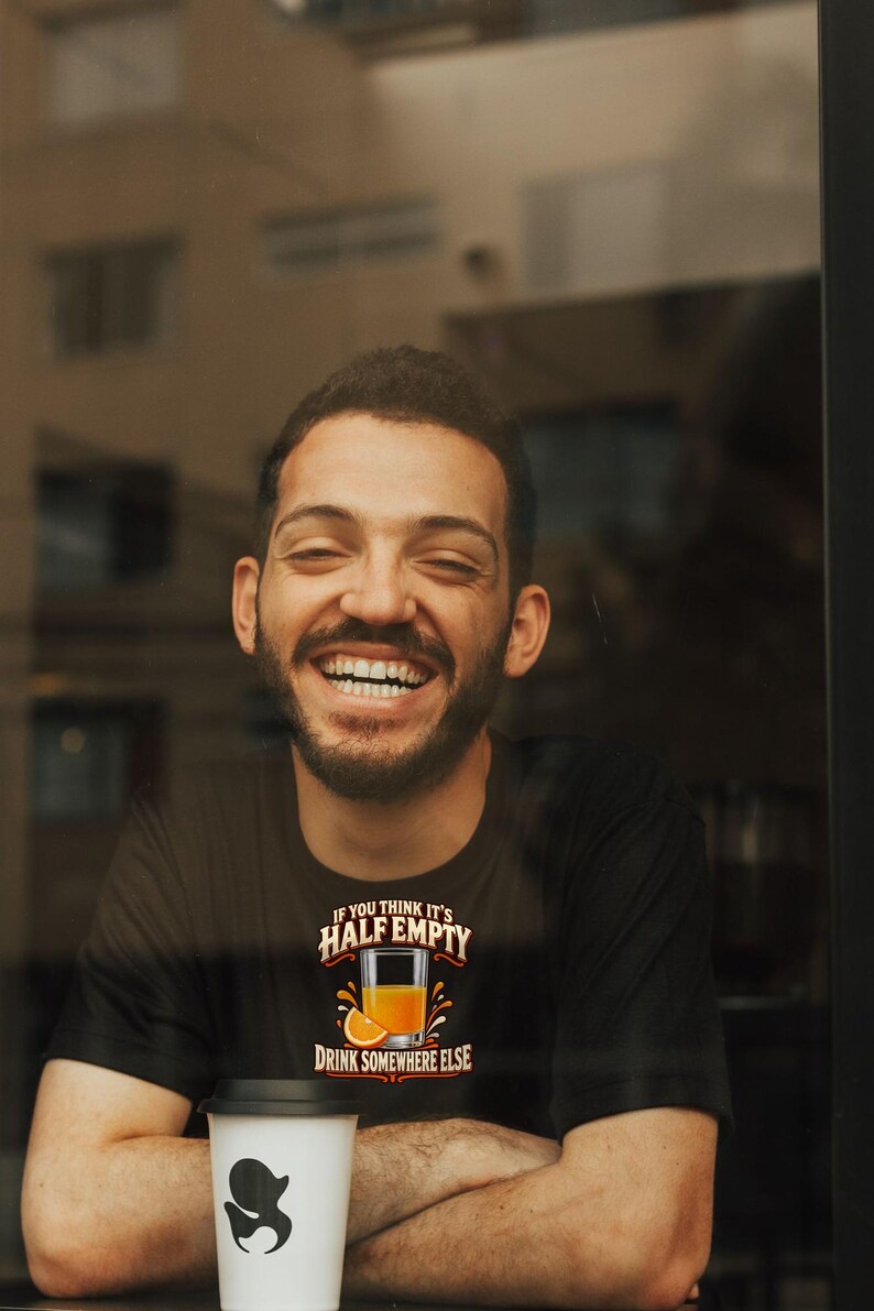 May include: A black t-shirt featuring a graphic of a glass of orange juice with an orange slice and the text IF YOU THINK IT'S HALF EMPTY DRINK SOMEWHERE ELSE. The shirt is worn with a white cup featuring a black logo in the foreground.