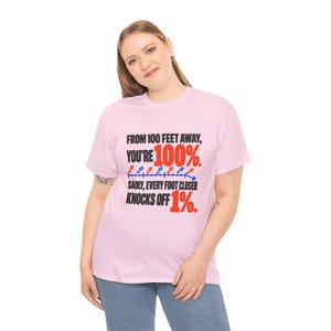 May include: A light pink short-sleeve t-shirt featuring black and red text that reads: FROM 100 FEET AWAY, YOU'RE 100%. SADLY, EVERY FOOT CLOSER KNOCKS OFF 1%. A graphic of small blue and red footprints follows a line with a downward arrow.