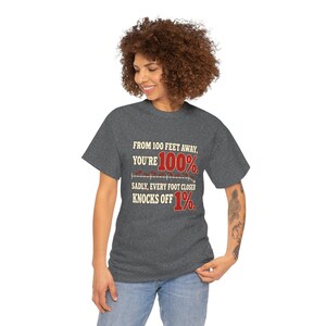 May include: A dark heather gray t-shirt featuring the text: FROM 100 FEET AWAY, YOU'RE 100%. SADLY, EVERY FOOT CLOSER KNOCKS OFF 1%. The design includes a red percentage graphic and a horizontal line with small red marks.