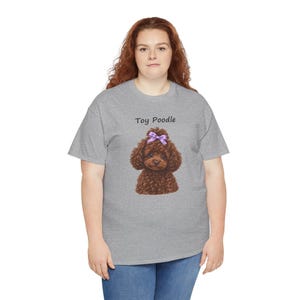 May include: A heather gray short-sleeve t-shirt featuring a graphic of a brown toy poodle with a purple bow on its head. The text Toy Poodle is printed in black above the dog illustration. The shirt has a crew neckline and a standard fit.