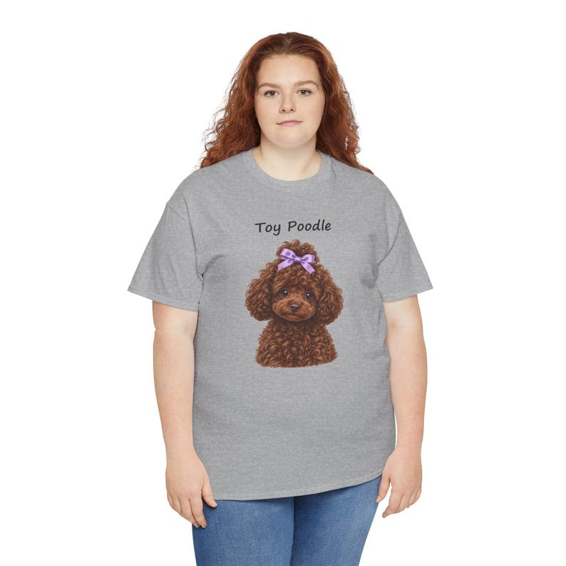 May include: A heather gray short-sleeve t-shirt featuring a graphic of a brown toy poodle with a purple bow on its head. The text Toy Poodle is printed in black above the dog illustration. The shirt has a crew neckline and a standard fit.