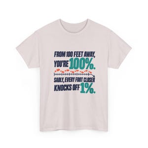 May include: A light pink short-sleeve t-shirt featuring dark blue and teal text that reads: FROM 100 FEET AWAY, YOU'RE 100%. SADLY, EVERY FOOT CLOSER KNOCKS OFF 1%. A graphic of a measuring line with orange footprints is centered.