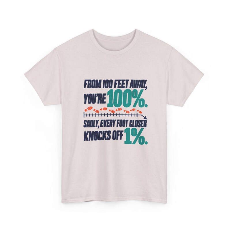 May include: A light pink short-sleeve t-shirt featuring dark blue and teal text that reads: FROM 100 FEET AWAY, YOU'RE 100%. SADLY, EVERY FOOT CLOSER KNOCKS OFF 1%. A graphic of a measuring line with orange footprints is centered.