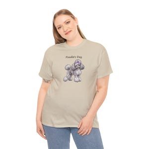 May include: A beige short-sleeve t-shirt featuring a graphic of a grey poodle wearing a purple bow on its head. Above the poodle, the text Poodle's Day is printed in a simple black font. The shirt has a crew neckline and a standard fit.