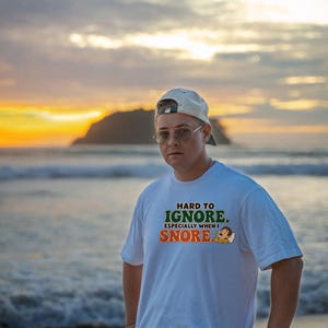 May include: A white short-sleeved t-shirt featuring the text "HARD TO IGNORE. ESPECIALLY WHEN I SNORE." in green and orange lettering, accompanied by a small graphic of a person sleeping on a pillow.