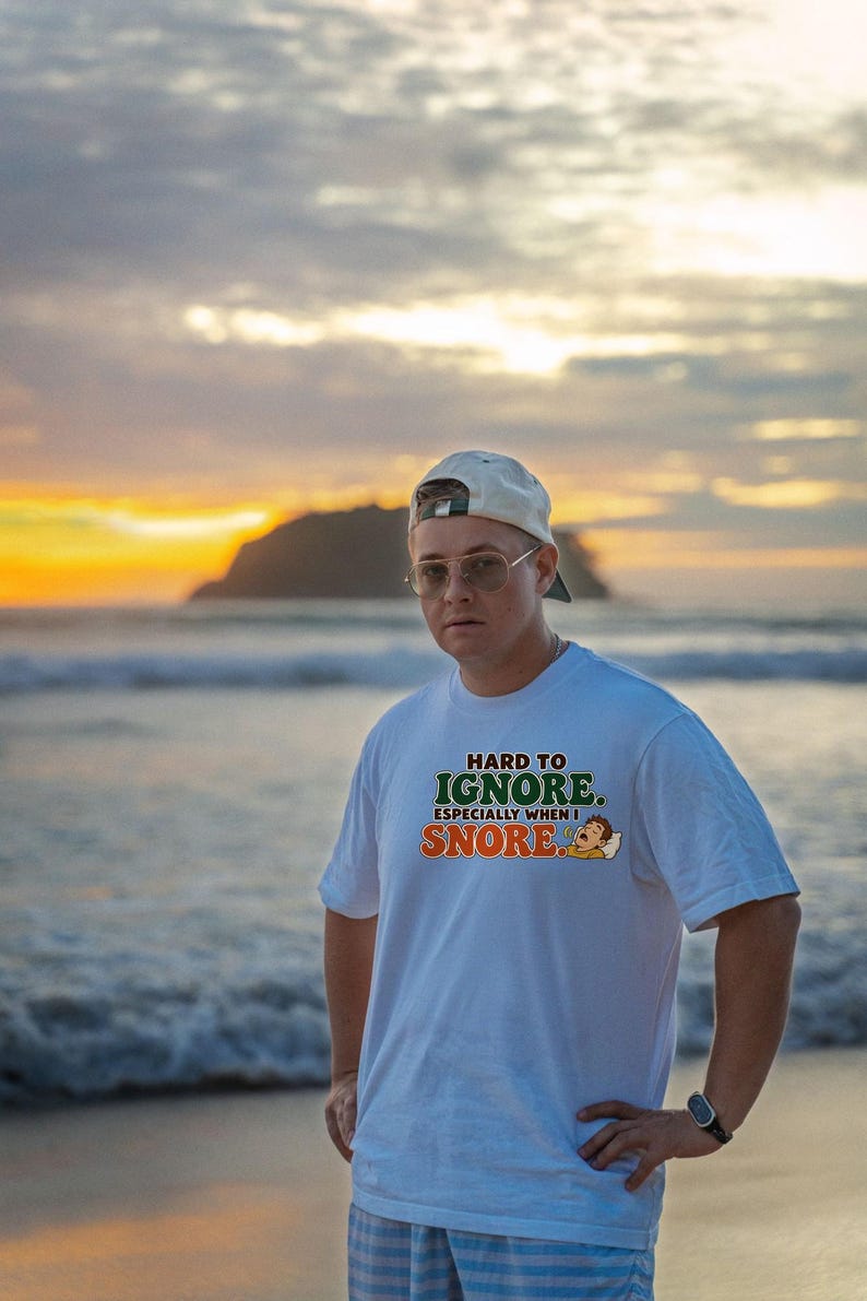 May include: A white short-sleeved t-shirt featuring the text "HARD TO IGNORE. ESPECIALLY WHEN I SNORE." in green and orange lettering, accompanied by a small graphic of a person sleeping on a pillow.