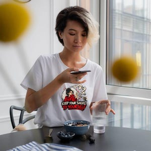 May include: A white short-sleeved t-shirt featuring a graphic print of a person in a red hoodie holding a hand up in a stop gesture. The text below the illustration reads KEEP YOUR SMOKE TO YOURSELF in bold red and black lettering.