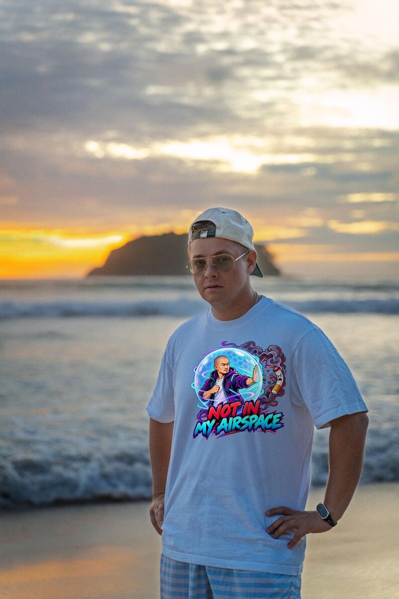 Może przedstawiać: Biały t-shirt z kr&oacute;tkim rękawem z nadrukiem graficznym przedstawiającym osobę w niebieskiej bańce energetycznej z napisem "NOT IN MY AIRSPACE" czerwonymi i niebieskimi literami, małą rakietą i fioletowym dymem.