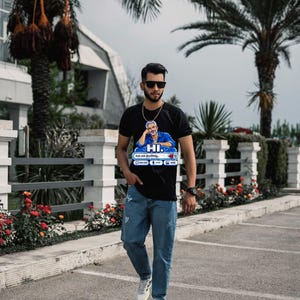 May include: A black short-sleeved t-shirt featuring a graphic of a person in a blue shirt with the text HI. Ask me anything... and icons for new chat, voice, and image. The shirt is paired with light blue distressed denim jeans and white trainers.
