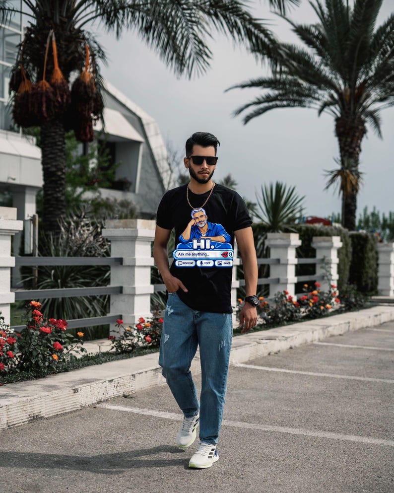 May include: A black short-sleeved t-shirt featuring a graphic of a person in a blue shirt with the text HI. Ask me anything... and icons for new chat, voice, and image. The shirt is paired with light blue distressed denim jeans and white trainers.