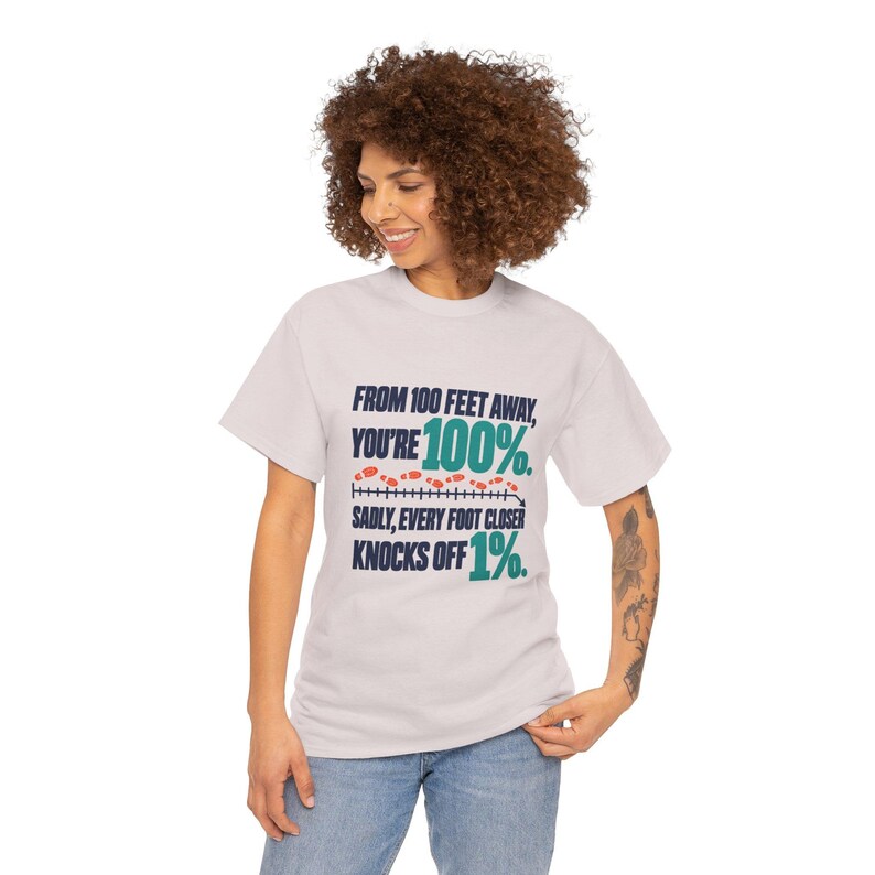 May include: A light gray short-sleeve t-shirt featuring dark blue and teal text that reads: FROM 100 FEET AWAY, YOU'RE 100%. SADLY, EVERY FOOT CLOSER KNOCKS OFF 1%. A graphic of a measuring line with small red footprints is included.
