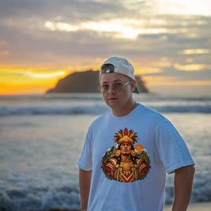 May include: A white short-sleeved t-shirt featuring a colorful graphic print of a figure wearing an elaborate feathered headdress and traditional gold-toned jewelry, set against a backdrop of stylized mountains and a sun.