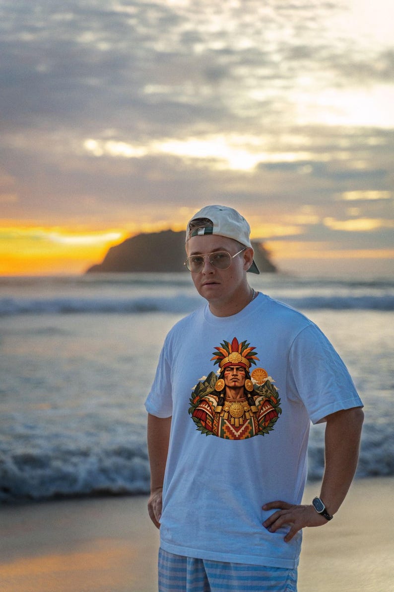 May include: A white short-sleeved t-shirt featuring a colorful graphic print of a figure wearing an elaborate feathered headdress and traditional gold-toned jewelry, set against a backdrop of stylized mountains and a sun.