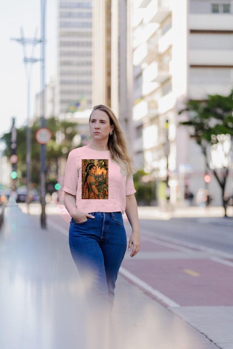May include: A light pink short-sleeved t-shirt featuring a rectangular graphic print of a person wearing traditional attire and jewelry, standing in a lush outdoor setting with trees and foliage under a warm, golden sunset sky.
